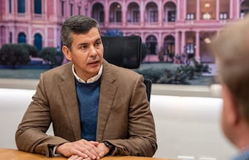 Santiago Peña Palacios sentado con chaqueta marrón y suéter azul, mientras un hombre escucha atentamente en un ambiente de oficina.