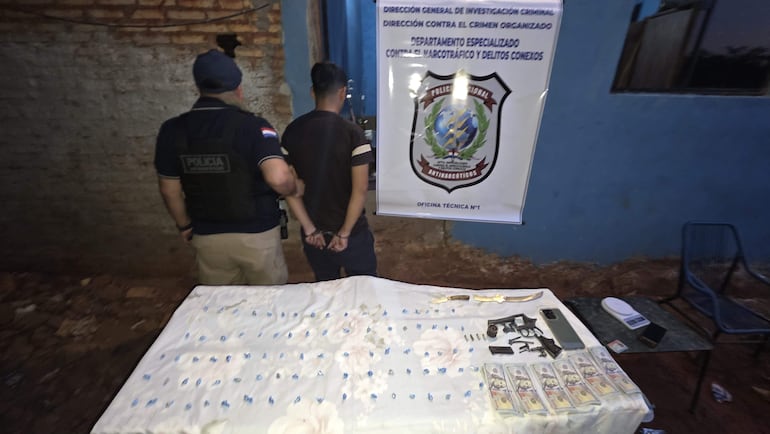 Policía presente junto a Jordan Larroza esposado, objetos como un arma y balanza sobre mesa blanca en ambiente azul claro.