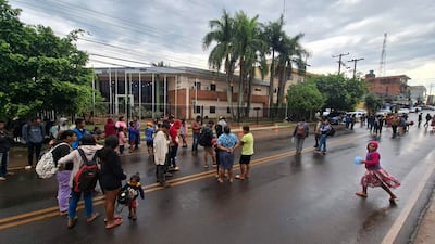 Grupo de personas de comunidades indígenas, algunos niños, en la calle frente a un edificio, con clima nublado y pavimento mojado.