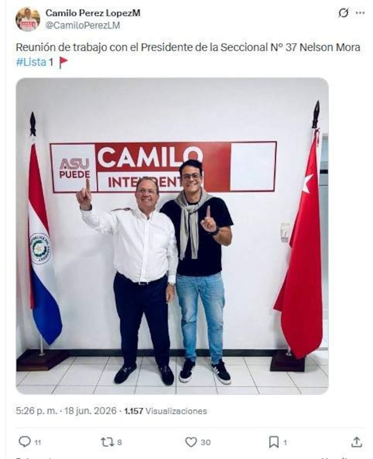 Camilo Pérez con camisa blanca y Nelson Mora en camiseta negra, ambos sonríen y hacen gestos de apoyo en un entorno de oficina.