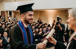 Joven graduado en toga y birrete sonríe mientras recibe su diploma de una mujer de cabello canoso, rodeados de otros graduados.