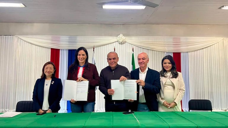 Claudia Centurión sonríe con blazer oscuro, mientras Basilio Núñez sostiene un documento en un evento con banderas de Paraguay.