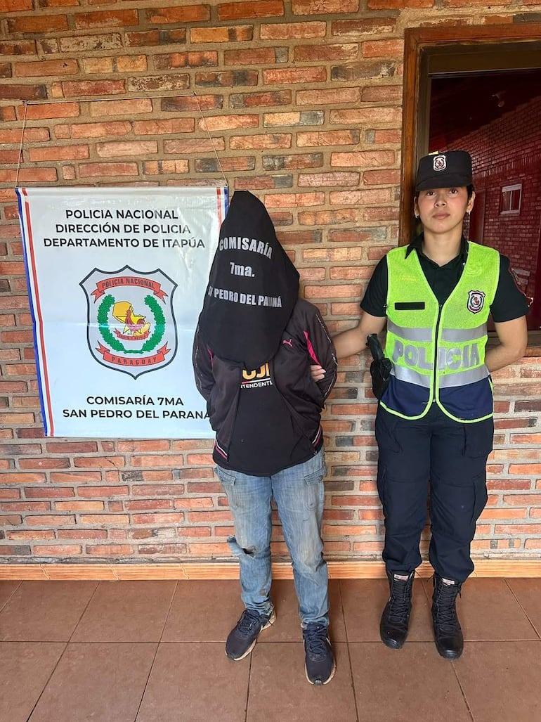 Individuo encapuchado acompañado por policía en comisaría, con un cartel que indica dirección de la Policía Nacional.