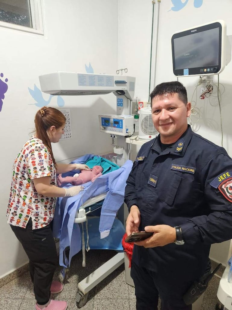 Mujer en uniforme de trabajo atiende a un recién nacido mientras un hombre con uniforme policial sonríe sosteniendo su teléfono.