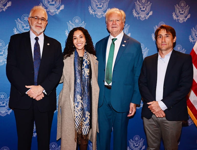 Hombre mayor con barba canosa y gafas, mujer joven en abrigo claro, hombre de mediana edad en traje azul, y hombre con saco oscuro en ambiente de conmemoración.