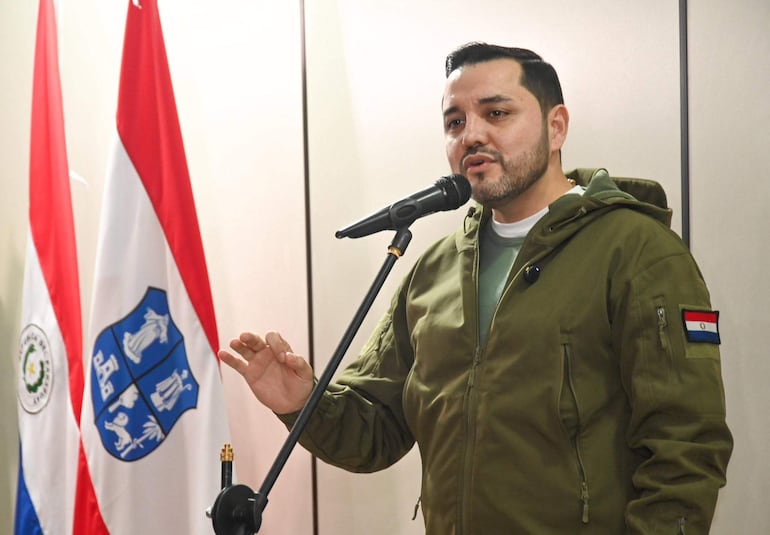 Luis Bello, en abrigo verde y suéter gris, gesticula frente a un micrófono, acompañado de banderas de Paraguay.