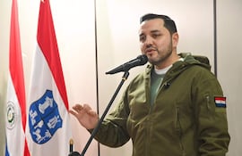 Luis Bello, en abrigo verde y suéter gris, gesticula frente a un micrófono, acompañado de banderas de Paraguay.