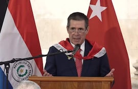 Orador en podio con traje oscuro y capa roja, banderas de Paraguay presentes en evento ceremonial.