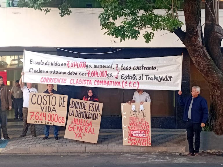 Cuatro personas protestan, dos sosteniendo un banner blanco y otros carteles, en un ambiente urbano. Todos lucen serios y decididos.