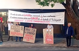 Cuatro personas protestan, dos sosteniendo un banner blanco y otros carteles, en un ambiente urbano. Todos lucen serios y decididos.