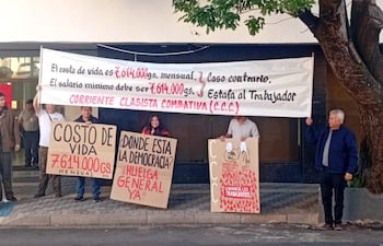 Cuatro personas protestan, dos sosteniendo un banner blanco y otros carteles, en un ambiente urbano. Todos lucen serios y decididos.