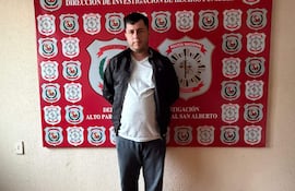 Hombre de pie en sala institucional, vestido con camiseta blanca, abrigo negro y pantalones grises. Fondo con escudos de la Policía Nacional.