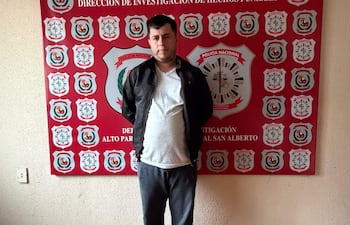 Hombre de pie en sala institucional, vestido con camiseta blanca, abrigo negro y pantalones grises. Fondo con escudos de la Policía Nacional.