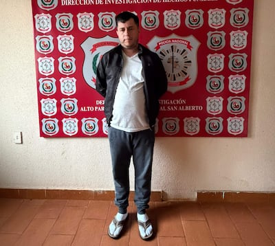 Hombre de pie en sala institucional, vestido con camiseta blanca, abrigo negro y pantalones grises. Fondo con escudos de la Policía Nacional.