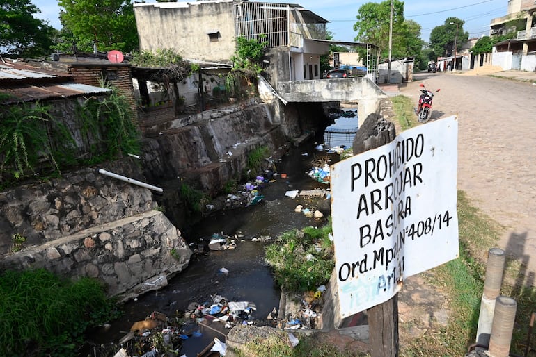 Cartel de 'PROHIBIDO ARROJAR BASURA' junto a un canal de aguas residuales lleno de desechos en un entorno urbano.