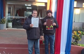 Arnaldo Aquino sostiene un documento frente a una institución pública, mientras un hombre a su lado sostiene un cartel y ambos visten abrigos.