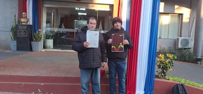 Arnaldo Aquino sostiene un documento frente a una institución pública, mientras un hombre a su lado sostiene un cartel y ambos visten abrigos.