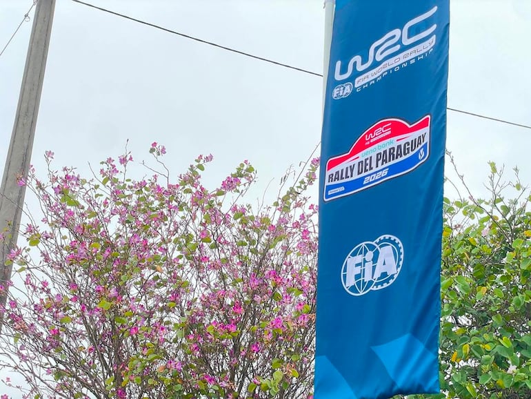 Banner colorido del Rally del Paraguay 2026, en un entorno urbano con arbustos floridos y cielo nublado.