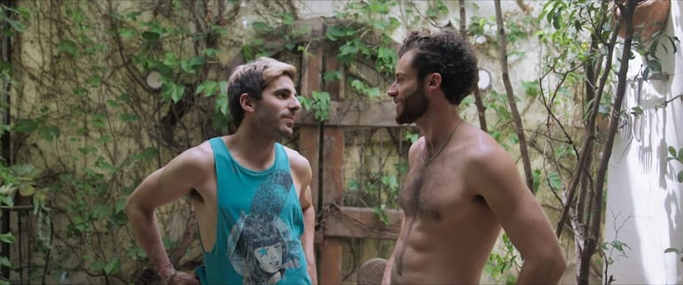 Dos hombres conversan en medio de vegetación, uno con camiseta azul y el otro sin camisa, en un ambiente íntimo.