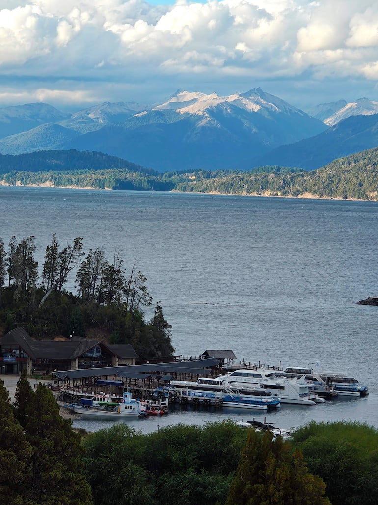 Muelle con embarcaciones amarradas, montañas nevadas al fondo y cielo parcialmente nublado en un ambiente sereno.