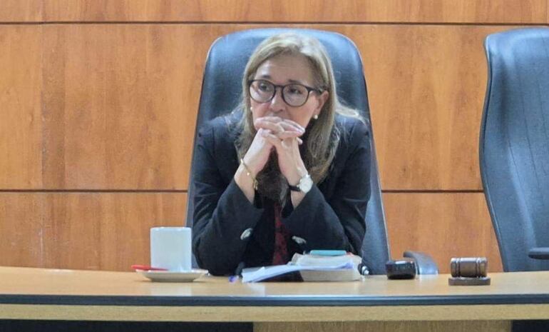 Jueza Mesalina Fernández con cabello largo y lacio, gafas, y blazer oscuro, sentada y reflexionando en una mesa con papeles.