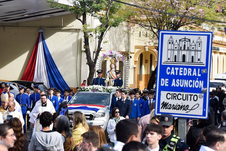 Grupo de hombres en uniforme flanquean a una figura religiosa sobre un vehículo, mientras un sacerdote dirige la ceremonia en un ambiente urbano.