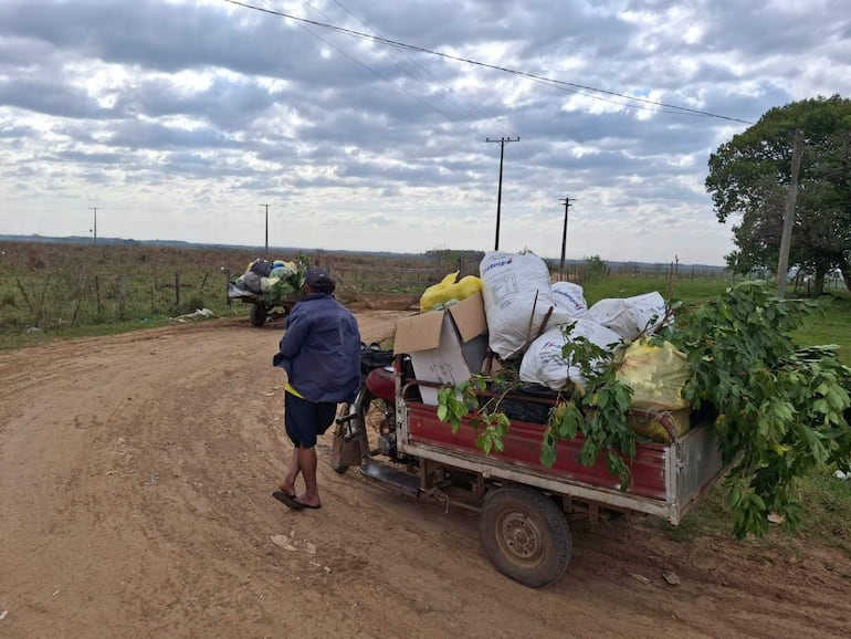 Hombre descalzo con camiseta amarilla y chaqueta oscura, junto a un motocarro cargado en un paisaje rural nublado.
