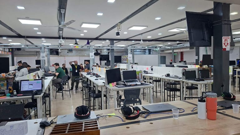 Espacio de trabajo moderno con aproximadamente diez personas, conversando y trabajando en computadores, ambiente dinámico y colaborativo.