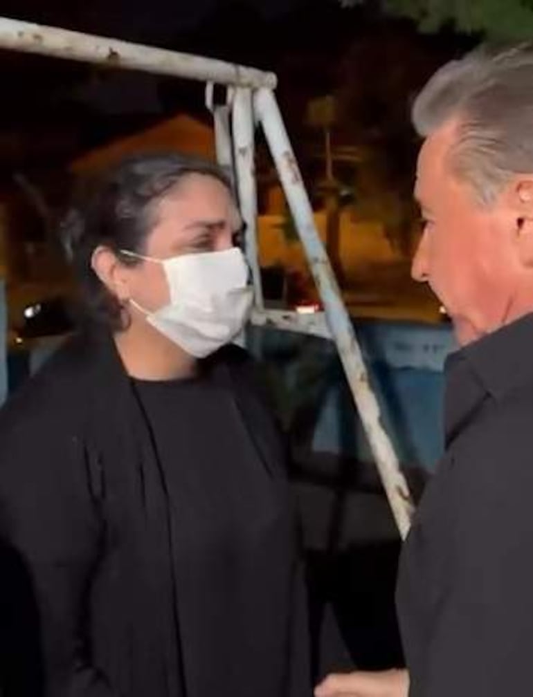 Mujer con máscara negra y hombre canoso en conversación, ambos vestidos de oscuro en un entorno iluminado.