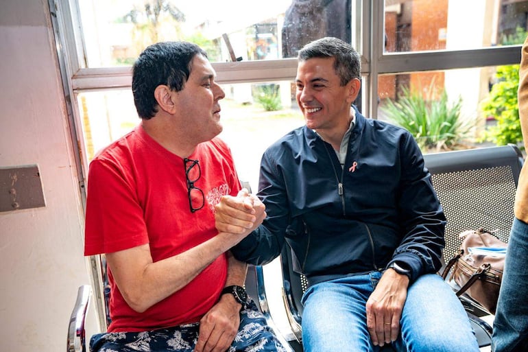 Santiago Peña en chaqueta oscura sonríe mientras conversa con un hombre de camiseta roja y pantalones estampados en un ambiente iluminado.