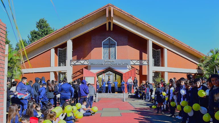 Grupo de niños con atuendos azules sostiene globos amarillos y blancos en la entrada de la parroquia.