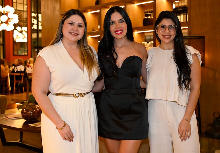 Tres mujeres sonrientes en vestidos formales, en un elegante salón con iluminación cálida y decoraciones de madera.