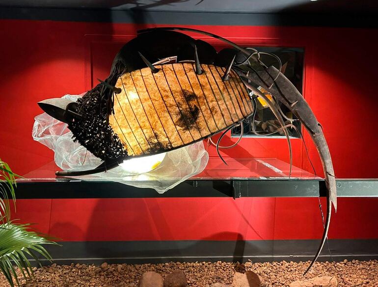 Obra artística de un pez metálico en vitrina con base de piedras en un ambiente cerrado iluminado.