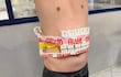 Individuo de pie con torso desnudo, sosteniendo un paquete de cigarrillos envuelto en cinta roja que dice 'FRÁGIL'.