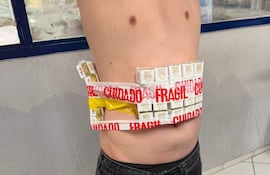 Individuo de pie con torso desnudo, sosteniendo un paquete de cigarrillos envuelto en cinta roja que dice 'FRÁGIL'.