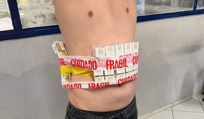 Individuo de pie con torso desnudo, sosteniendo un paquete de cigarrillos envuelto en cinta roja que dice 'FRÁGIL'.