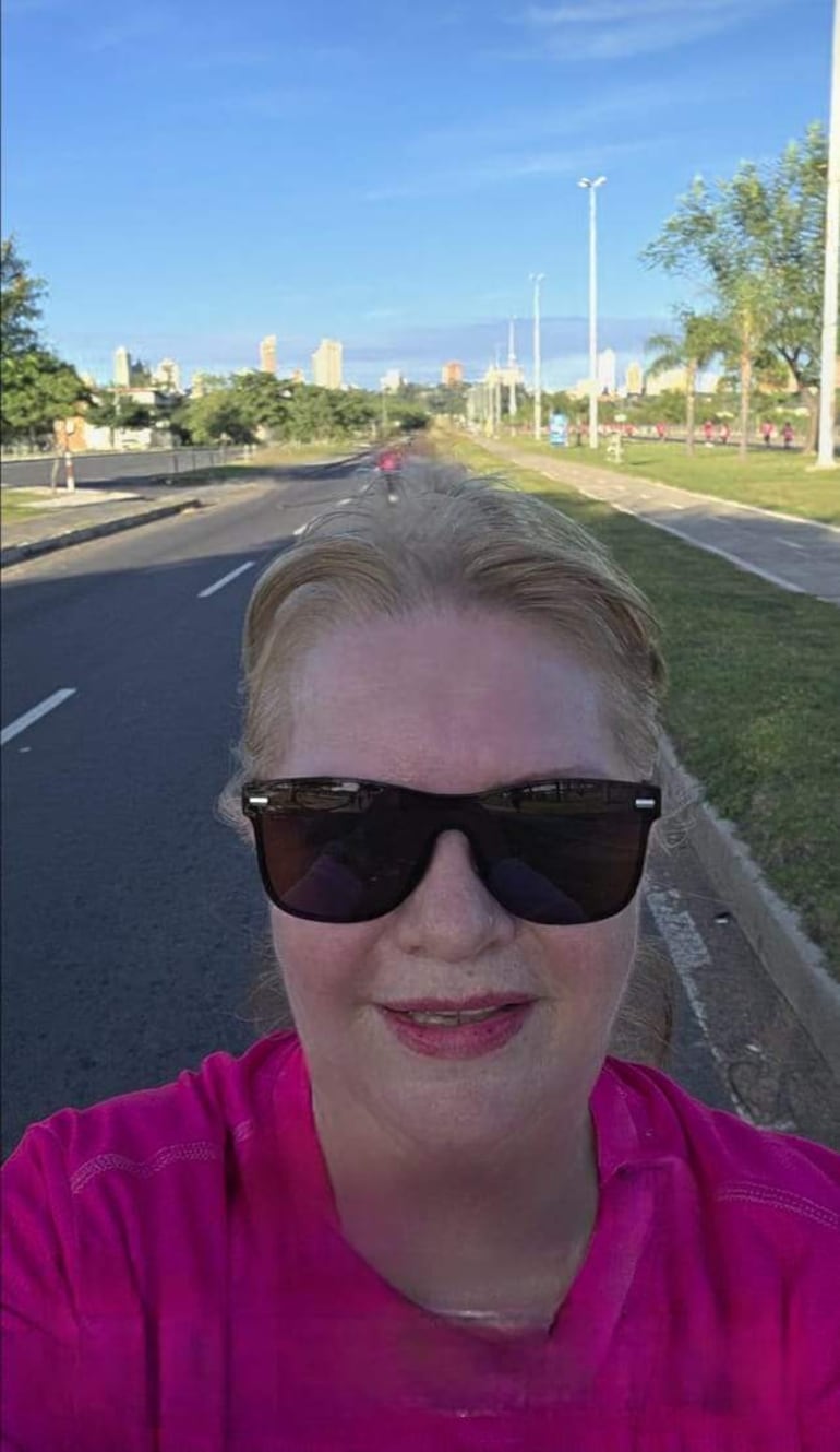 Mujer de cabello claro en camiseta rosa y gafas de sol, sonríe en un ambiente recreativo con personas al fondo.