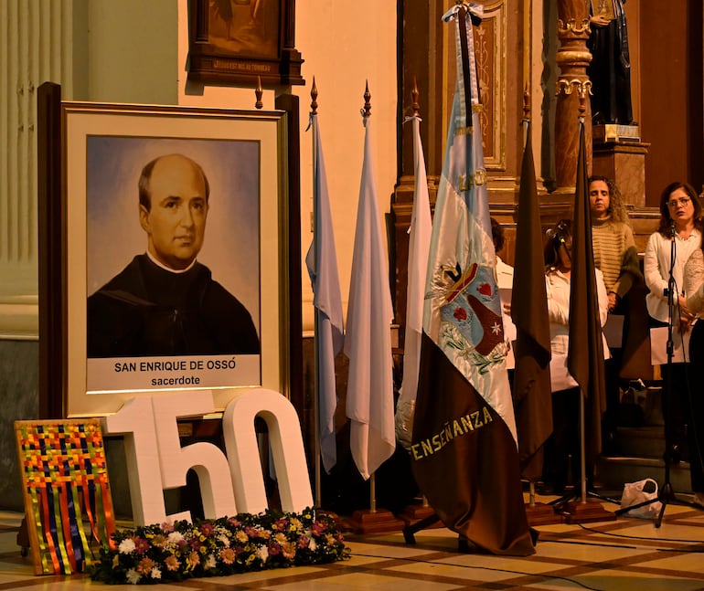 Retrato de San Enrique de Ossó en el altar, personas atentas escuchando, banderas blancas y azules decoran el ambiente solemne.