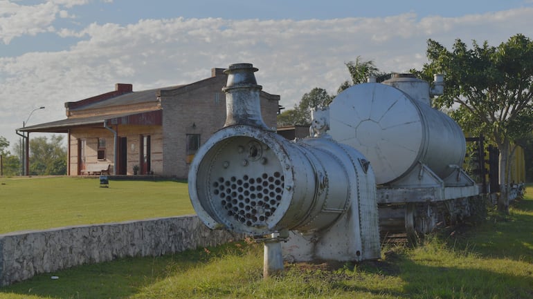 Bosque verde alrededor de una antigua locomotora y un edificio colonial de ladrillo, bajo un cielo nublado.