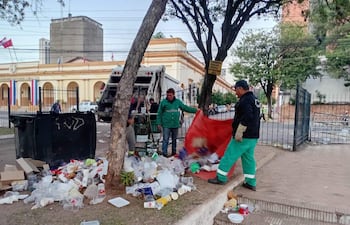 Tres trabajadores en uniformes verde y negro recogen basura en una vereda frente a un edificio institucional.