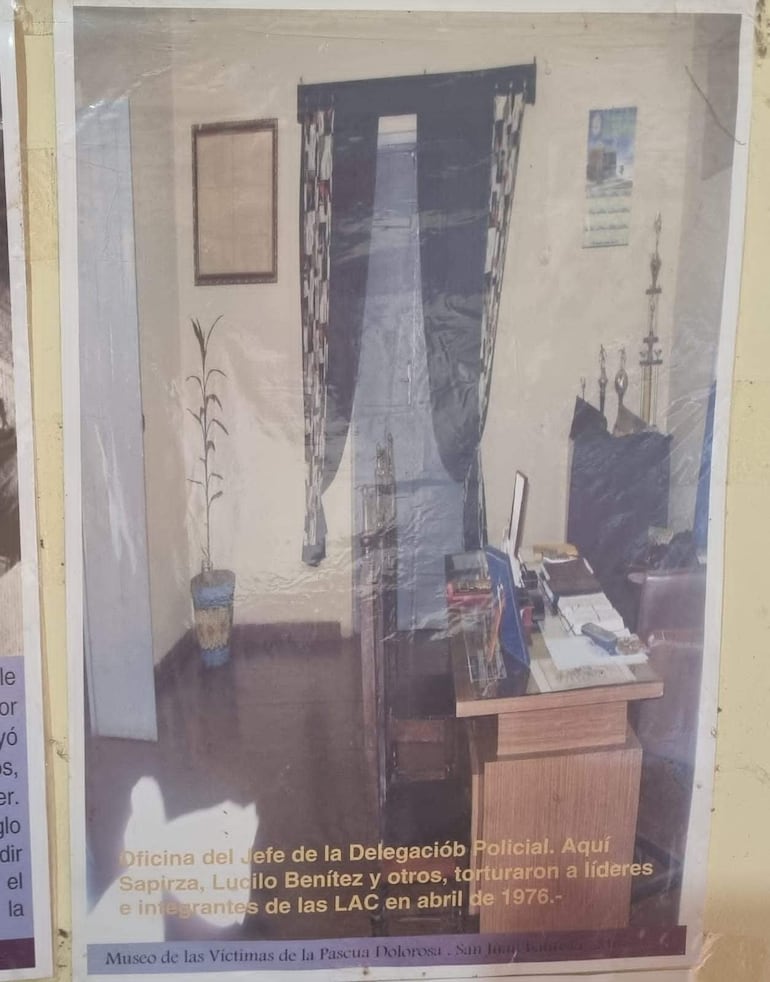 Oficina austera con escritorio de madera, teléfono y documentos. Cortinas geométricas oscuras y una planta decorativa en la esquina.