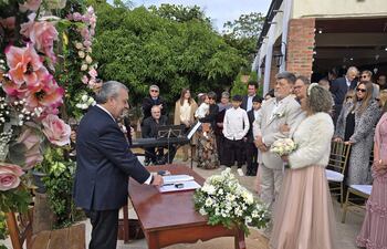 Ricardo Flecha de traje oscuro habla a la audiencia, mientras Fanny Pereira sostiene un ramo de flores, rodeados de decoraciones florales.