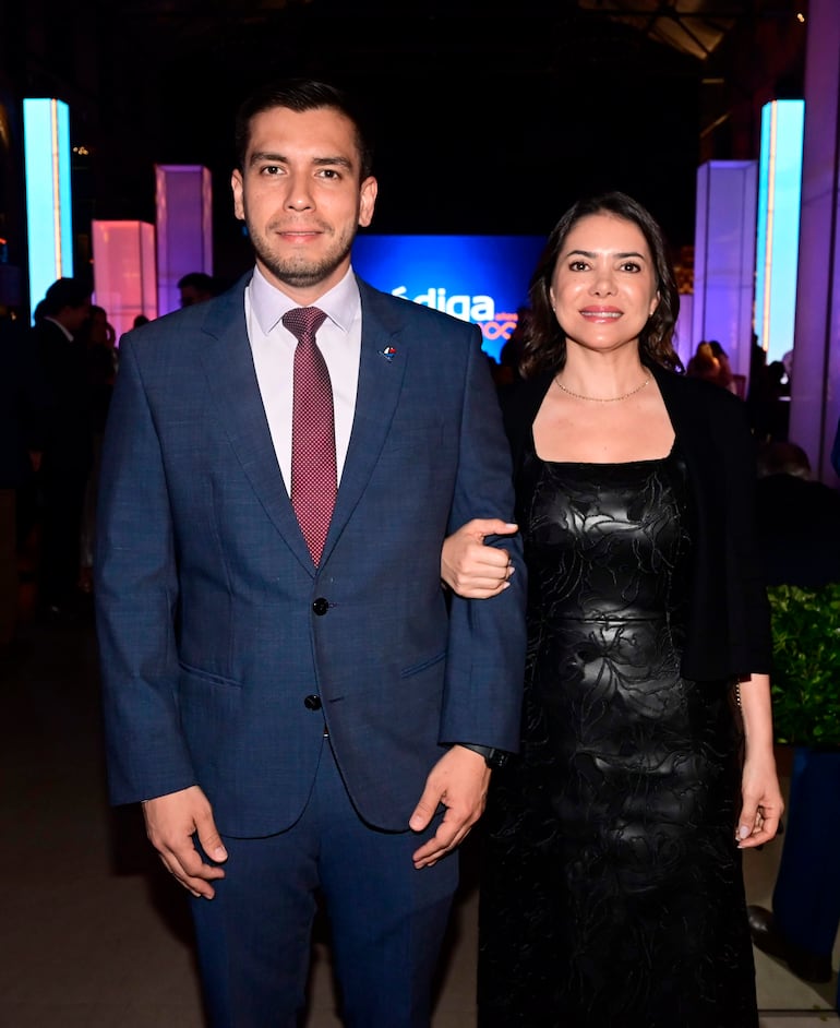 Mujer en vestido negro y hombre en traje azul sonríen en evento formal, con pantallas iluminadas de fondo.
