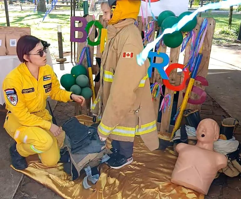 Mujer con uniforme amarillo agachada hablando con un niño de pie vestido de bombero, decoraciones coloridas al fondo.