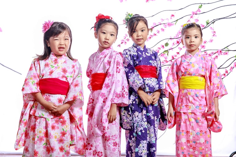 Cuatro niñas con kimonos coloridos, posando en un fondo blanco decorado con flores artificiales, luciendo sonrisas tranquilas.