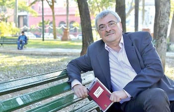 Hombre canoso con gafas, vestido con saco oscuro y camisa de rayas, sentado en banco leyendo, rodeado de árboles y una plaza.