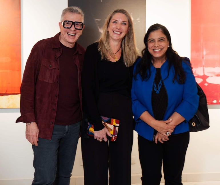 Tres personas sonríen en un espacio cultural, una mujer con abrigo azul, dos en atuendos oscuros, rodeados de arte abstracto.