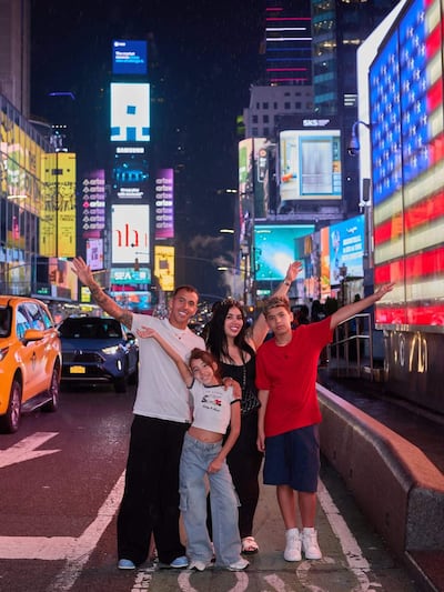 Alejandro Romero Gamarra Kaku, mujer con cabello oscuro y niños en camiseta blanca y roja, posan felices bajo pantallas LED en Times Square.