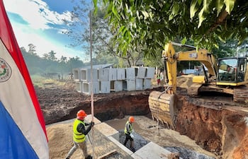 Cuatro trabajadores en casco y chalecos reflectantes en obra, con excavadora y bloques de cemento. Bandera de Paraguay visible.
