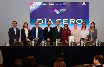 Elías Saba en conferencia, rodeado de personas de pie con expresiones tranquilas, en un salón decorado con sobriedad.